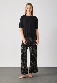 Crna majica kratkih rukava uparena s crnim širokim hlačama s cvjetnim uzorkom i zlatnim naglascima, na neutralnoj pozadini.