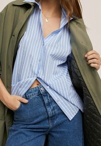 Chemise à rayures bleues et blanches, à col, à manches longues, rentrée dans un jean bleu taille haute. Manteau vert olive drapé sur les épaules.