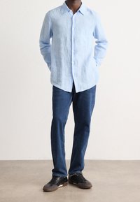 Camicia in lino azzurro chiaro con colletto, bottoni sul davanti e tasca sul petto, abbinata a jeans di denim blu scuro e sneakers nere.