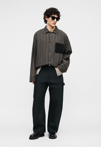 Άνδρας με μαύρα γυαλιά ηλίου, φαρδιά ριγέ πουκάμισο με κουμπιά και υφασμένη τσέπη, φαρδιά μαύρα παντελόνια και μαύρα παπούτσια, στέκεται μπροστά σε λευκό φόντο.