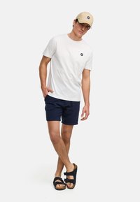 Witte katoenen t-shirt met een klein marineblauw logo, gecombineerd met marineblauwe textuurschoenen en zwarte sandalen met twee gespen. Pet in beige.