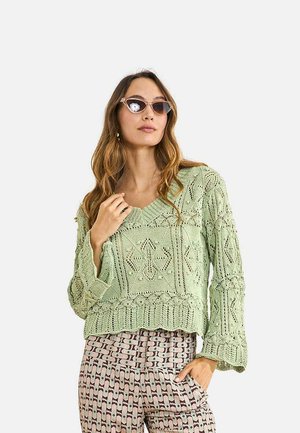 Mujer con suéter de punto color verde claro con un patrón de diamantes y pantalones de talle alto estampados, complementada con gafas de sol de estilo ojo de gato.