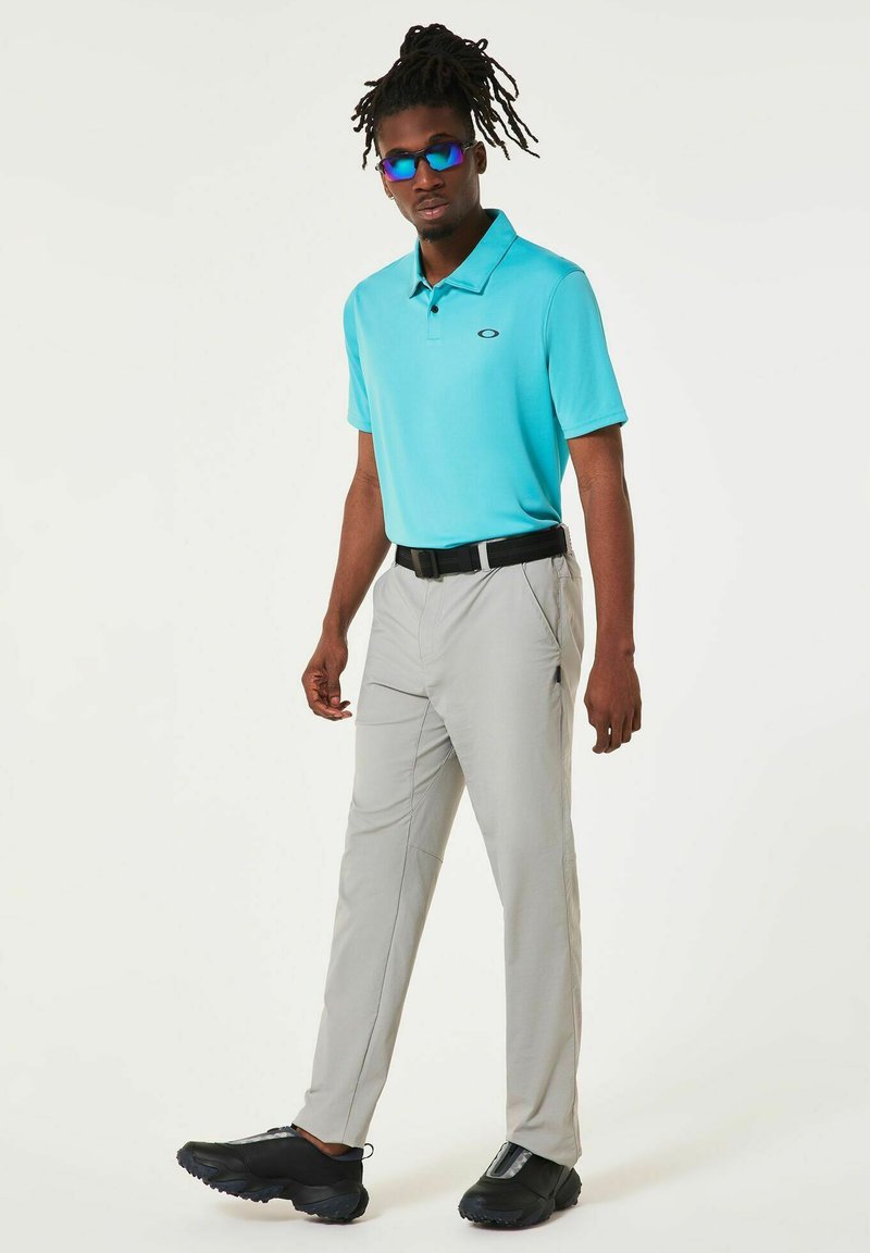 Polo bleu clair à manches courtes, pantalon gris, ceinture noire et baskets noires avec semelles texturées ; le modèle porte des lunettes de soleil bleues.