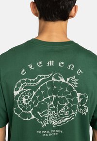 Maglietta verde scuro con una grande grafica bianca di un rettile stilizzato con contorni bold sul retro, il testo sottostante recita: "CREEP, CRAWL, OR ROLL."