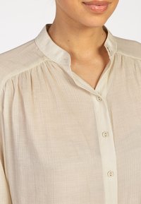 Beige button-up shirt made of lightweight, textured fabric. Features a stand collar and gathered detailing at the shoulders.