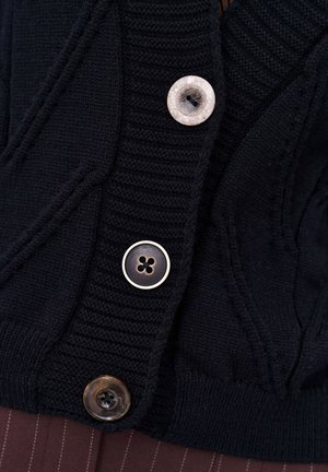 Black knit cardigan featuring ribbed texture, three buttons in various styles: white, gold, and dark brown with a four-hole design.