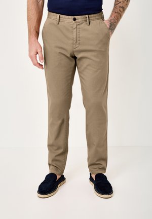 Man wearing beige chinos with front pockets, a navy shirt, navy espadrille shoes, and visible arm tattoos.