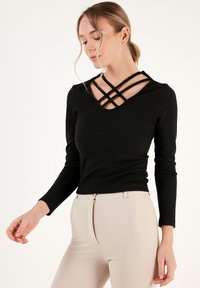 Top lungo a maniche nere a coste con scollo a V e cinghie incrociate. Abbinato a pantaloni beige aderenti con dettaglio della zip.