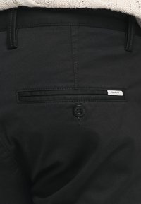 Schwarze Hose mit glatter Oberfläche, ausgestattet mit einer Gesäßtasche, einem Knopfverschluss und einem kleinen GANT-Logo-Tag.