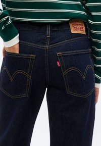 Άτομο που φοράει σκούρο μπλε τζιν Levi's με καφέ ραφές και πράσινο-λευκό ριγέ μακρυμάνικο πουκάμισο, με το χέρι στην πίσω τσέπη.