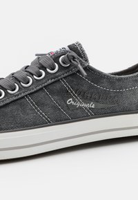 Gray canvas sneaker with white rubber sole, featuring silver eyelets, gray laces, and logo detail. Stitching provides additional texture.