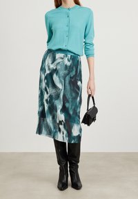 Cardigan turquoise avec boutons associé à une jupe plissée à mi-mollet avec un motif abstrait bleu et blanc. Bottes noires montantes et sac à main.