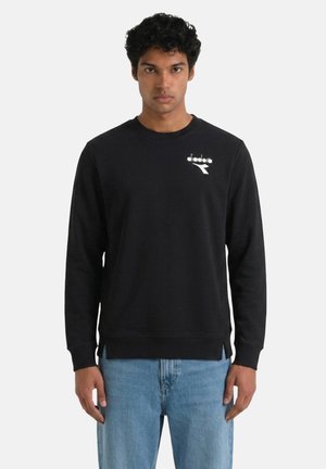 Uomo che indossa una felpa nera a maniche lunghe con un piccolo logo bianco Diadora sul petto e jeans azzurri, in piedi davanti a uno sfondo bianco.
