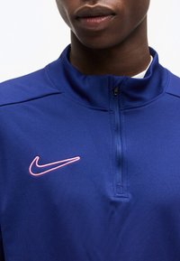 Κοντινό πλάνο ενός ατόμου που φορά μπλε αθλητικό τοπ με φερμουάρ και ροζ λογότυπο Nike στο αριστερό στήθος.