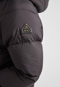 Czarna ocieplana kurtka z rękawem, na którym znajduje się trójkątny czarny i metaliczny naszywka z logo "JACKIT" na górnej części ramienia, na jednolitym tle.
