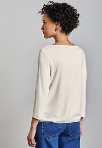 Lichtbeige, gestructureerde langwerpige top met een wijde halslijn, gecombineerd met hoge blauwe denim broeken. Minimalistisch ontwerp, geen patronen.
