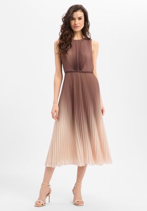 Frau in einem ärmellosen, plissierten Midikleid mit einem Ombre-Verlauf von Braun zu Beige, trägt nudefarbene High Heels vor weißem Hintergrund.