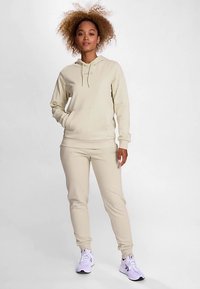 Crèmekleurige hoodie en bijpassende joggingbroek gemaakt van zachte stof; voorzien van een voorzak, capuchon met trekkoord en elastische boorden; gecombineerd met witte sneakers.