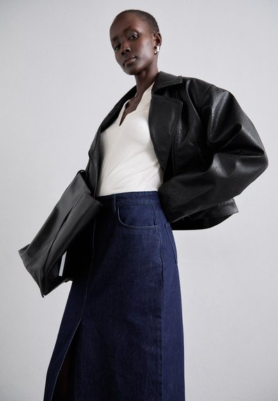 Zwarte leren cropped jas, wit V-hals shirt en lange donkere denim rok met een zijsplit; model houdt een zwarte handtas vast.