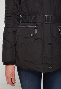 Svart vadderad jacka med bälte i midjan, dragkedje- och tryckknappsfickor, samt kviltad textur med en slät yta.