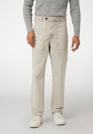 Beige Canvas-Hosen mit entspanntem Schnitt, ausgestattet mit Seitentaschen und einem Knopfverschluss. Kombiniert mit weißen Sneakern.