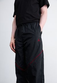 Zwarte joggerbroek met een trekkoord in de taille, voorzien van rode accenten en een logo aan de linkerkant. Lichtgewicht stof met een zachte textuur.