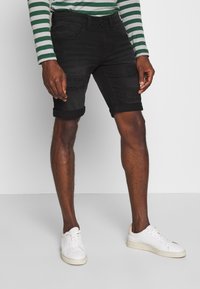 Shorts in denim nero con dettagli strappati, risvolti e cinque tasche, abbinati a sneakers bianche e una camicia a maniche lunghe con righe verdi.