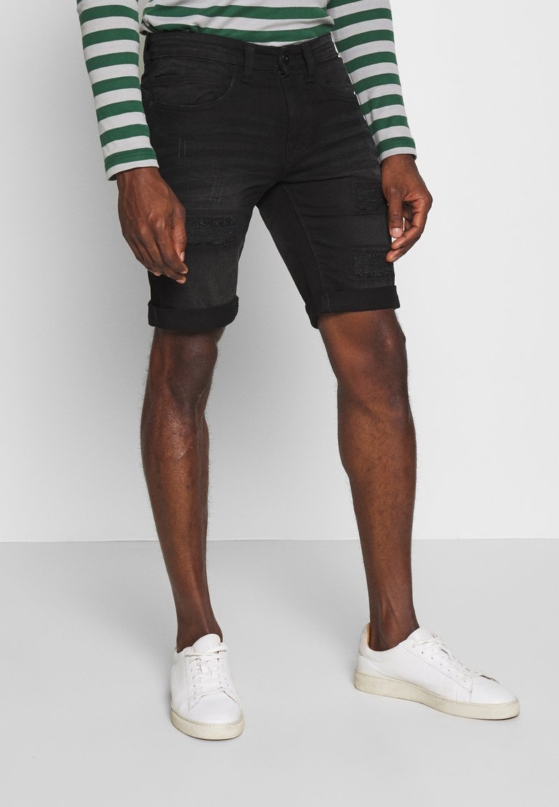 Shorts in denim nero con dettagli strappati, risvolti e cinque tasche, abbinati a sneakers bianche e una camicia a maniche lunghe con righe verdi.