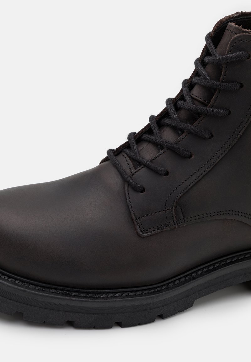 Brązowy skórzany but o gładkim wykończeniu, okrągłym nosku i czarnych sznurówkach. Chropowata gumowa podeszwa z głębokim bieżnikiem zapewnia dobrą przyczepność.