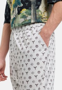 Pantalons blancs à motifs avec des designs géométriques marron. Ils sont dotés d'une taille élastique et d'une poche sur le côté. Tissu lisse avec une coupe décontractée.
