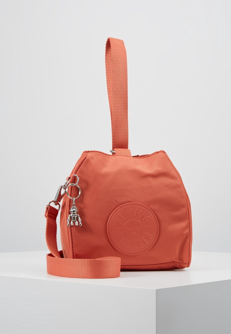 Mała torba z koralowego materiału z okrągłym logo Kipling, odczepiany pasek na ramię, uchwyt na górze i srebrny charms w kształcie małpki.