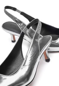 Zapatos de tacón metálicos plateados con un diseño elegante, que cuentan con una correa texturizada y un interior negro. Tacón bajo y punta en forma afilada.