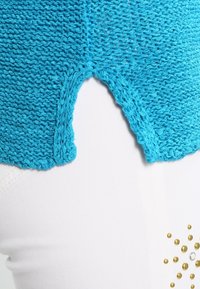 Haut tricoté turquoise avec une découpe en V au niveau de l'ourlet. Le tissu texturé contraste avec un pantalon blanc lisse orné de clous métalliques.