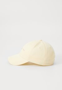Žltá baseballová čiapka s mierne zakriveným okrajom, so zošitými ventilačnými otvormi a jemným písaním v ružovej farbe na prednej strane.