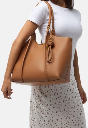 Brown leather tote bag with a smooth texture, featuring a woven handle and knotted accent. The bag is spacious with a structured shape.