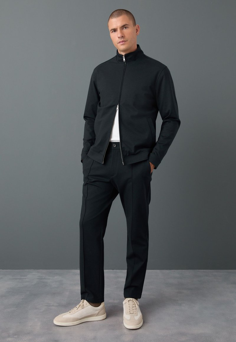 Mann mit schwarzer Zip-up-Jacke, schwarzer Hose, weißem Hemd und beigen Sneakers, steht mit den Händen in den Taschen vor einer grauen Wand.