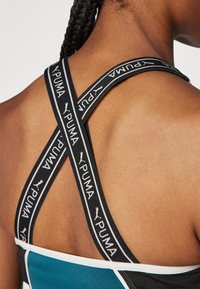 Ingrandimento della schiena di una persona che indossa una maglietta sportiva blu con fasce elastiche nere con logo Puma che si incrociano sulle spalle.