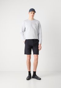 Šviesiai pilkas megztinis su apvaliu kaklu ir kvadratu, derinamas su juodomis šortais, juodomis kojinėmis ir tamsiomis loafer bateliais. Modelis stovi priešais vienspalvį foną.