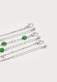 Pulseras de cadena de plata con motivos de ojos verdes y varios estilos de cierre, incluyendo detalles de garra de langosta y bola, dispuestas en una fila.