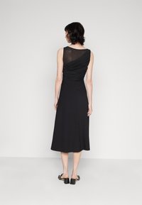 Femme portant une robe midi noire sans manches avec un tissu transparent froncé au dos supérieur, se tenant devant un fond clair.