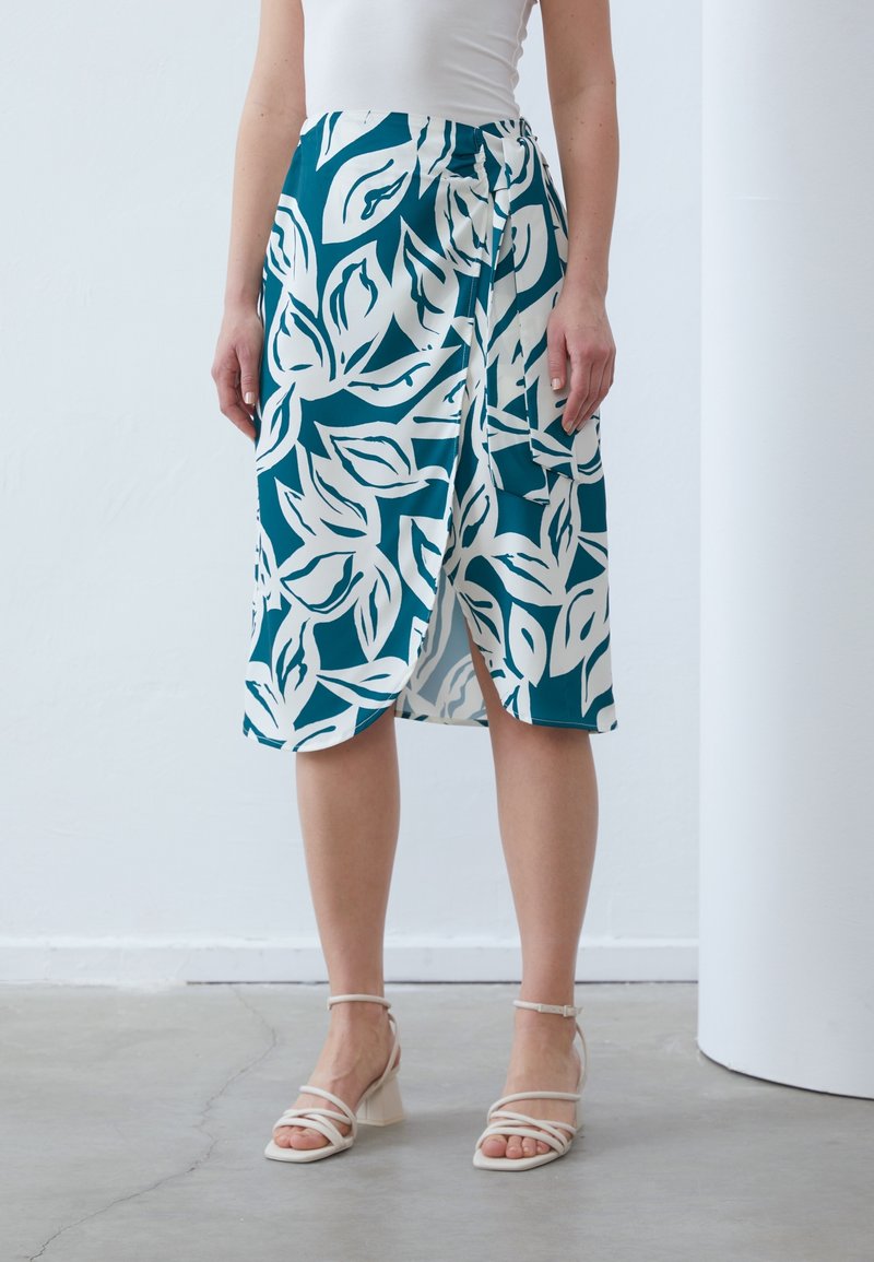 Femme portant des talons blancs à brides et une jupe mi-longue bleu sarcelle avec de grands motifs de feuilles blanches et une fente latérale, debout sur un sol en béton.