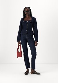 Giacca di jeans blu scuro con scollatura quadrata, chiusura a buttoni e due tasche frontali, abbinata a jeans coordinati e scarpe basse nere. Tiene in mano una borsa rossa.