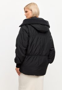 Zwarte pufferjack met een gestructureerde donkergrijze capuchon, elastische taille en manchetten. Beschikt over een losse, volumineuze draad en een zachte afwerking.