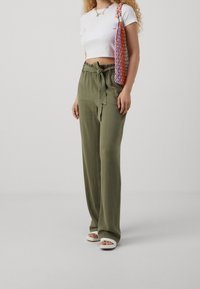Pantalon large couleur vert olive avec taille à nouer et texture douce, porté avec un top court blanc et des sandales légères à enfiler de couleur claire.
