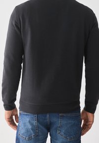 Felpa nera in cotone, con collo rotondo, polsini e orlo a coste. Indossata con jeans blu in denim con texture sbiadita.