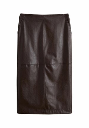 Frau trägt grünen gestreiften Pullover, langen schwarzen Rock, schwarze Absätze, große Sonnenbrille und hält eine schwarze Tasche, während sie sich an einen Pfosten lehnt.