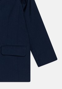 Mørkeblå skreddersydd jakke med en glatt tekstur, dekorert med en klafflomme, spisse slag og rene linjer i stoffkonstruksjonen.