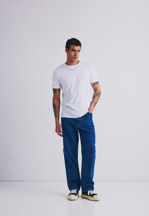Weißes T-Shirt mit kurzen Ärmeln, blaue locker sitzende Jeans, schwarz-weiße Sneaker, silberne Kettenhalskette, stehend vor einem einfarbigen Hintergrund.