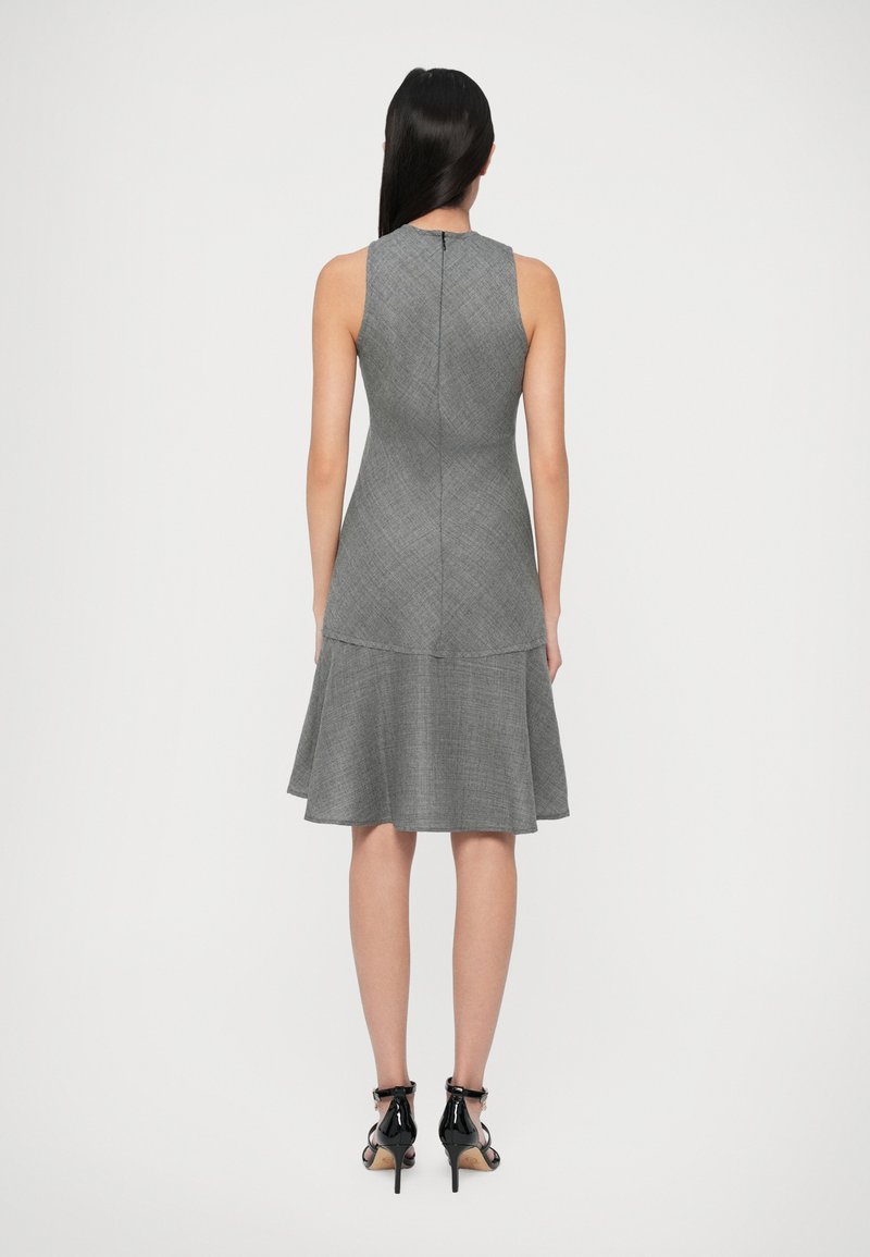 Femme aux longs cheveux noirs portant une robe grise sans manches arrivant aux genoux et des sandales à talons hauts noires, se tenant de dos sur un fond blanc.