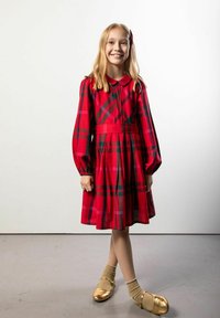 Młoda dziewczyna uśmiechająca się, ubrana w czerwoną sukienkę w kratę, złote buty z beżowymi skarpetkami, stojąca ze skrzyżowanymi nogami na jasnoszarej podłodze na białym tle.